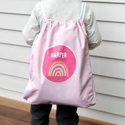 Child wearing pink drawstring swim bag with rainbow print and personalised name.