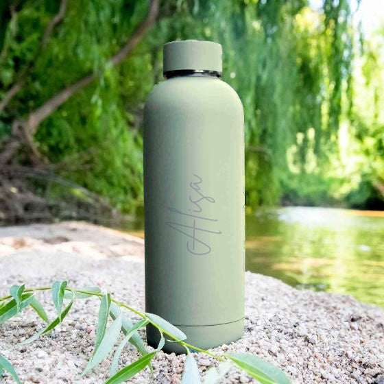 Personalised sage green drink bottle engraved with the name Alisa in script, standing on river pebbles beside a willow tree